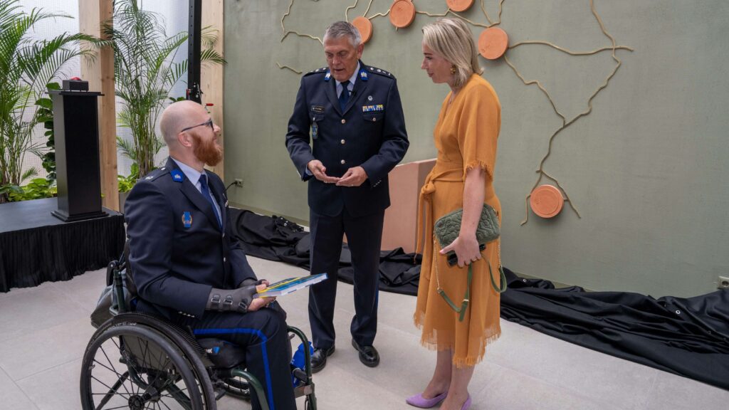 Foto met daarop van links naar rechts: auteur Ralph Stoové, DV&O directeur Erik de Borst en voormalig staatssecretaris Ingrid Coenradie. Stoové en De Borst dragen als uniform het ceremonieel tenue en Coenradie draagt een mosterdgele jurk. Ralph zit in een rolstoel.