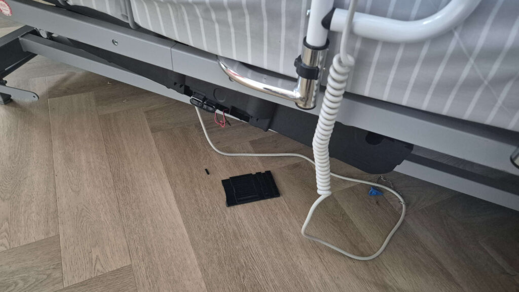 Afbeelding van een stukje verzorgingsbed van Medipoint met een afgebroken en loshangende bedmotor en losse afgebroken onderdelen onder het bed.