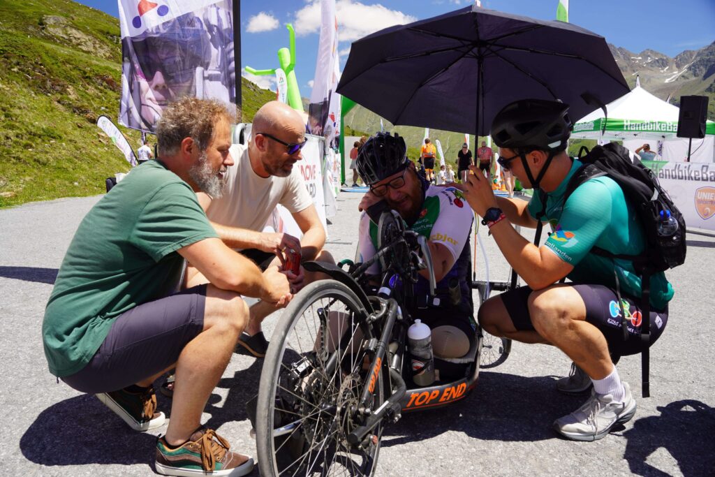 Prachtig emotioneel moment na de finish van de Handbike Battle van Ralph Stoové. Ralph zit nog in zijn zwarte handbike met rode letters Top End Force 3. Ralph huilt. Links van hem knielen vrienden Jaap en Rob en rechts knielt buddy Jonas. Ze zorgen voor troost en support na deze beladen overwinning. Ralph draagt een zwarte korte fietsbroek en een turquoise met groen en wit fietsshirt, oftewel het officiële deelnemersshirt van de HandbikeBattle. Ralph heeft links een hoge bovenbeenamputatie en rechts heeft hij zijn verlamde been in een zwarte enkelvoetorthese. Aan zijn handen draagt hij polshand-orthesen. Hij draagt ook steunkousen.