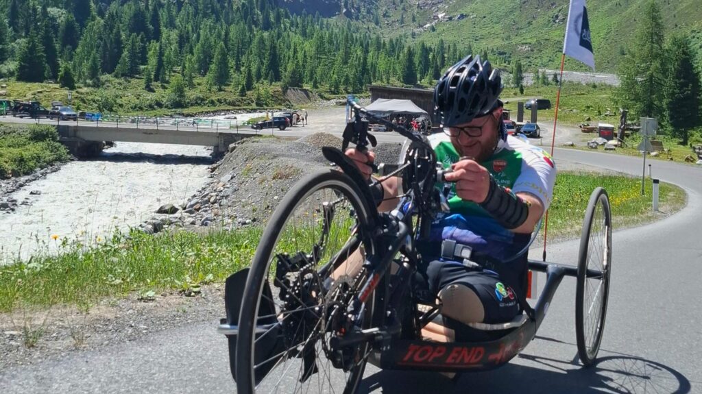 Foto van handbiker Ralph Stoové tijdens de Handbike Battle 2026. Ralph fietst op een zwarte handbike met rode letter Top End Force 3. Ralph heeft links een hoge bovenbeenamputatie en rechts heeft hij zijn verlamde been in een zwarte enkelvoetorthese. Ralph draagt een zwarte korte fietsbroek en een turquoise met groen en wit fietsshirt, oftewel het officiële deelnemersshirt van de HandbikeBattle. Hij draagt ook steunkousen. Op de achtergrond zie je een bergachtig landschap met een bergrivier en een brug. Je ziet heel duidelijk het hoogteverschil.