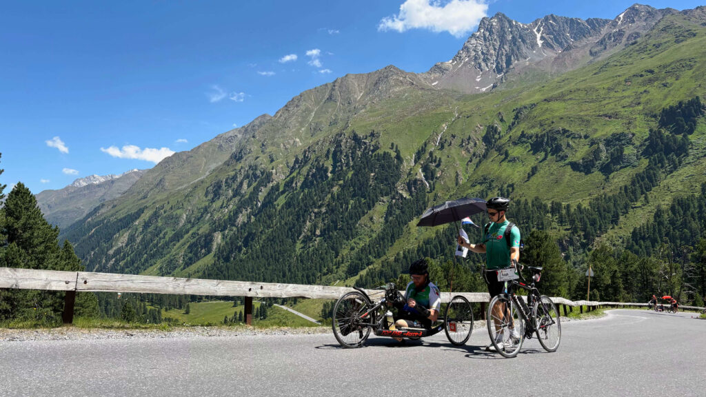 Foto van Ralph Stoové met zijn buddy Jonas tijdens de Handbike Battle 2026. Ralph draagt een zwarte korte fietsbroek en een turquoise met groen en wit fietsshirt, oftewel het officiële deelnemersshirt van de HandbikeBattle. Hij draagt ook steunkousen. Op de achtergrond zie je een bergachtig landschap. Jonas loopt met zijn racefiets aan de hand naast de handbiker en heeft een paraplu vast om Ralph tegen te zon te beschermen. Jonas draagt een mintgroen fietsshirt van team Birds on Wheels.