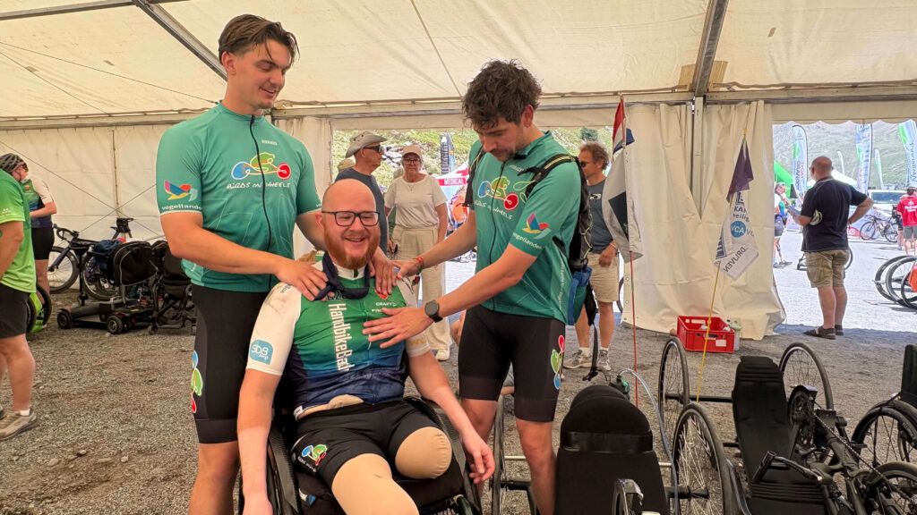 Foto van Ralph Stoové in zijn rolstoel na de finish van de Handbike Battle 2026. Ralph ziet er zeer vermoeid en uitgeput uit. Ralph moet door twee buddy's overeind gehouden worden. De buddy's dragen het mintgroene teamshirt van Birds on Wheels en Ralph draagt het turquoise, groen met witte wedstrijdshirt van de HandbikeBattle. Ralph heeft links een hoge bovenbeenamputatie met een stompkousje om zijn stomp en rechts een steunkous om zijn verlamde been. Je ziet een stukje van zijn stomazakje boven zijn zwarte fietsbroek uitkomen.