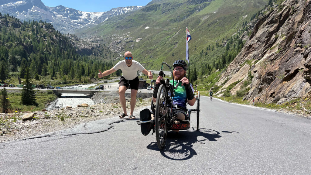 foto van handbiker Ralph Stoové tijdens de Handbike Battle 2026. Ralph wordt enthousiast aangemoedigd door een van zijn vrienden. Ralph fiets op een zwarte handbike met rode letter Top End Force 3. Ralph heeft links een hoge bovenbeenamputatie en rechts heeft hij zijn verlamde been in een zwarte enkelvoetorthese. Ralph draagt een zwarte korte fietsbroek en een turquoise met groen en wit fietsshirt, oftewel het officiële deelnemersshirt van de HandbikeBattle. Hij draagt ook steunkousen. Op de achtergrond zie je een bergachtig landschap met een bergrivier en een brug. Je ziet heel duidelijk het hoogteverschil.